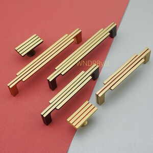 Puede incluir: Una colecci&oacute;n de herrajes dorados para gabinetes, que incluyen manijas y perillas. Las manijas presentan un dise&ntilde;o lineal con tres barras paralelas y extremos rectangulares. Las perillas tienen un dise&ntilde;o similar. El hardware se muestra sobre una superficie roja y gris.