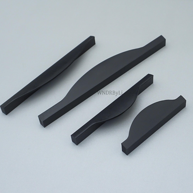 May include: Four matte black cabinet pulls in varying sizes. The pulls have a curved, ergonomic design and a rectangular profile. The hardware is displayed on a light gray background. The pulls are likely for drawers or cabinets.