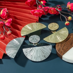 Pode incluir: Uma coleção de ferragens decorativas para armários em acabamentos dourado e prateado. As ferragens incluem puxadores semicirculares e puxadores redondos com um design de raios de sol. Flores vermelhas e um leque vermelho ao fundo. O texto "WNDbyLi" é visível.