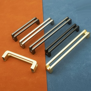 May include: A selection of cabinet pulls in gold, silver, black, and gunmetal finishes. The pulls have a modern, minimalist double-bar design. The hardware is displayed against a contrasting orange and blue background. Measurements are in centimetres.