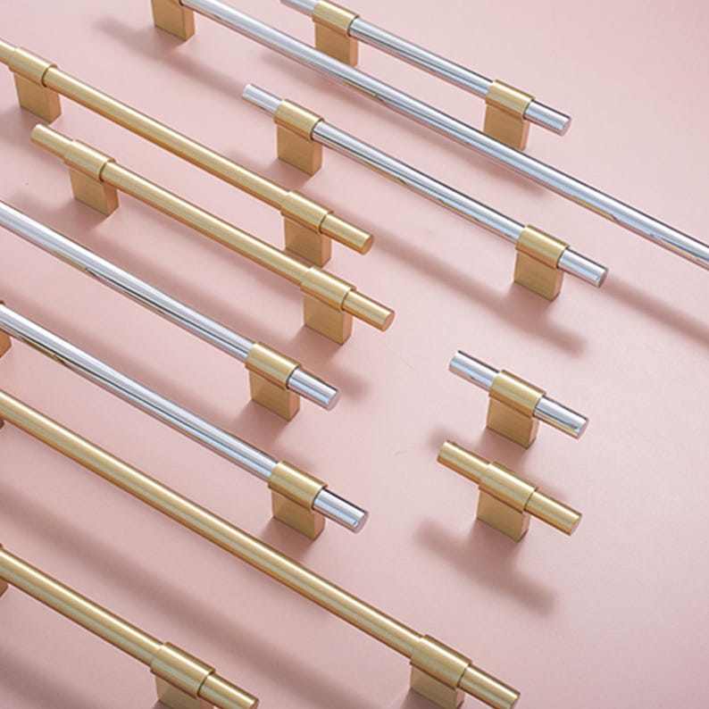 Op de afbeelding: Een assortiment kastgrepen in goud en zilver. De T-vormige handgrepen zijn gemonteerd op rechthoekige bases. De hardware wordt gepresenteerd op een roze oppervlak, waarbij de verschillende stijlen voor interieurontwerp worden benadrukt.