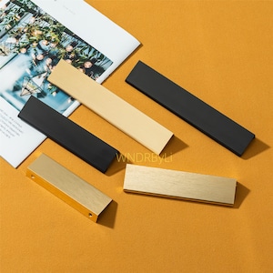 Op de afbeelding: Een assortiment kastgrepen in goud, zwart en lichte houttinten is gerangschikt op een mosterdkleurig oppervlak. De grepen zijn rechthoekig, met een verscheidenheid aan afwerkingen. Een tijdschrift met een geometrisch ontwerp staat op de achtergrond.