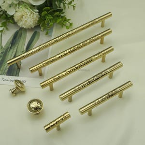 May include: A collection of gold-colored cabinet hardware, including several bar pulls and two knob styles. The pulls have a textured, hammered design, and the knobs feature a similar hammered texture. The hardware is arranged on a light-colored surface.