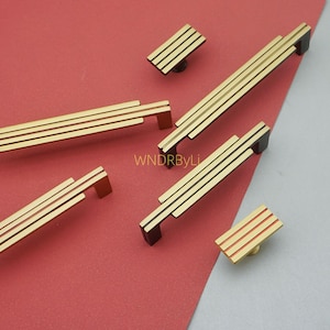 Puede incluir: Juego de herrajes dorados para armarios con un dise&ntilde;o moderno. El juego incluye manijas y pomos de varios tama&ntilde;os, con un dise&ntilde;o lineal de tres rayas. Los herrajes tienen detalles en negro y se muestran sobre un fondo rojo y gris.