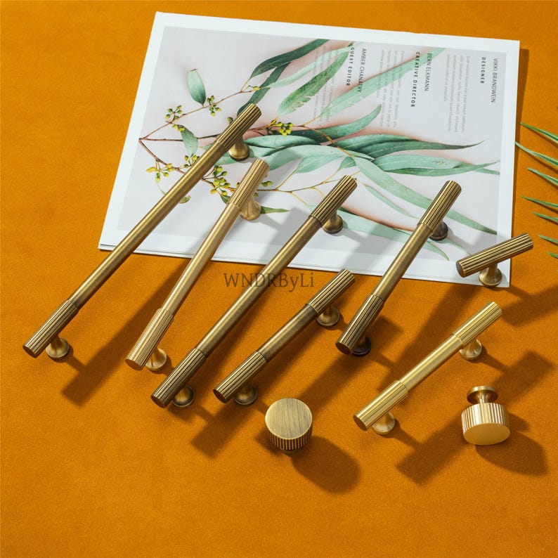 Puede incluir: Una colecci&oacute;n de herrajes de lat&oacute;n para gabinetes, que incluye manijas y perillas, dispuestas sobre una superficie. El hardware presenta un dise&ntilde;o acanalado y un color dorado c&aacute;lido. La imagen incluye una revista con texto y vegetaci&oacute;n.