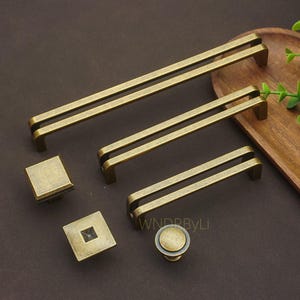 May include: A selection of antique brass cabinet hardware, featuring square and round knobs, alongside double-bar pull handles. The hardware has a vintage, aged look, with a warm, golden-brown colour. The handles are available in different lengths.