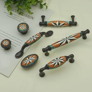 以下が含まれることがあります： 装飾的なキャビネット金具のコレクション。ノブとプルが含まれています。 金具は、白、オレンジ、ターコイズのスターバーストデザインが特徴で、茶色の背景にあります。 金具は金属製です。