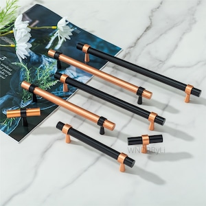 May include: A collection of cabinet pulls in black and copper finishes. The pulls have a cylindrical bar with copper accents and are arranged on a marble surface. The text "WNDRByLi" is visible on the right side of the image.