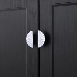 May include: Close-up of a pair of cabinet door handles. Each handle features a white, fan-shaped design with a black base. The handles are mounted on a dark gray wooden cabinet door, creating a contrast in color and texture.