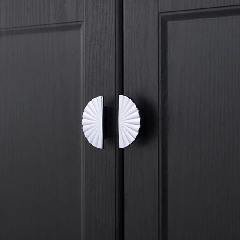 May include: Close-up of a pair of cabinet door handles. Each handle features a white, fan-shaped design with a black base. The handles are mounted on a dark gray wooden cabinet door, creating a contrast in color and texture.