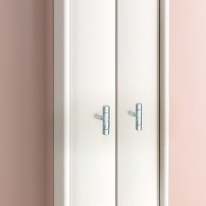 May include: White cabinet doors with two silver-colored, textured T-shaped handles. The cabinet doors are set against a soft pink wall. The handles appear to be made of metal.