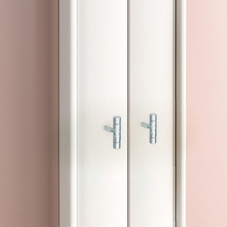 May include: White cabinet doors with two silver-colored, textured T-shaped handles. The cabinet doors are set against a soft pink wall. The handles appear to be made of metal.