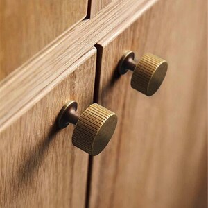 Puede incluir: Primer plano de dos pomos de armario de lat&oacute;n con un dise&ntilde;o estriado. Los pomos est&aacute;n montados en una puerta de armario de madera marr&oacute;n claro. Los pomos tienen forma circular y un acabado met&aacute;lico c&aacute;lido.