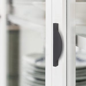 May include: A close-up of a black cabinet handle with a curved design. The handle is mounted on a white cabinet door. The background is blurred, showing a glimpse of white plates.