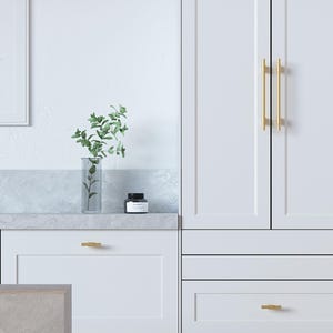 Op de afbeelding: Een helder, modern keukenontwerp met witte kasten en gouden hardware. Een glazen vaas met groen en een kleine zwarte pot staan op een marmeren aanrecht. De kastdeuren hebben verticale gouden handgrepen.