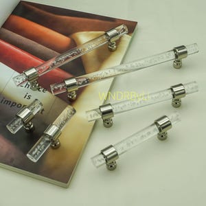 May include: Clear acrylic cabinet handles with silver-coloured fittings. The handles feature a bubble design within the acrylic. The handles are arranged on a book with text that reads "is important".