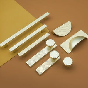 May include: A collection of gold-colored cabinet hardware, including bar pulls, half-moon pulls, and round knobs. The hardware features a ridged design and is arranged on a brown and yellow background.