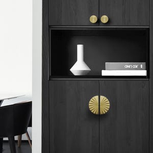 May include: Dark wood cabinet with gold-colored, sunburst-style knobs. The cabinet has a recessed shelf with a white vase and two books. The cabinet doors have a large, half-circle knob. The cabinet is in a modern style.