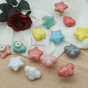 May include: A selection of colourful ceramic cabinet knobs in star and cloud designs. The knobs are in various colours, including pink, yellow, blue, and grey, with gold-coloured hardware. The text "WNDRByLi" is visible.