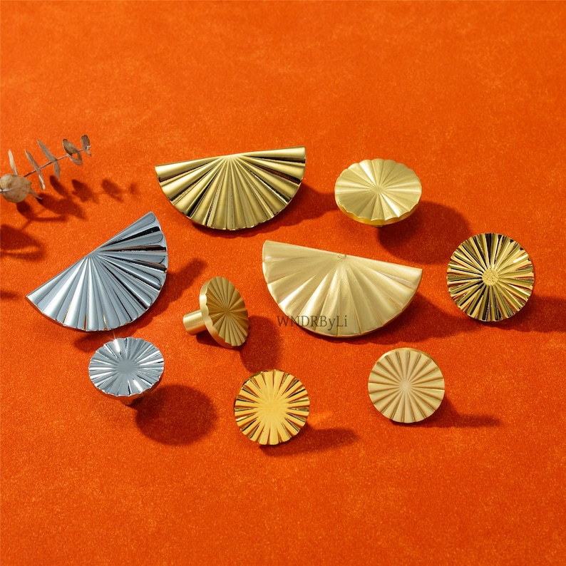 May include: An assortment of decorative metal hardware in gold and silver tones, including half-circle and circular shapes. The hardware features a fan-like design with radial grooves. The items are arranged on an orange surface.