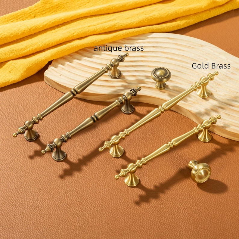 Peut inclure: Une s&eacute;lection de quincaillerie d'armoire en laiton antique et en laiton dor&eacute;, comprenant des poign&eacute;es et des boutons. Les poign&eacute;es sont allong&eacute;es avec des d&eacute;tails d&eacute;coratifs, tandis que les boutons sont ronds. La quincaillerie est pr&eacute;sent&eacute;e sur une surface en bois et une surface marron.