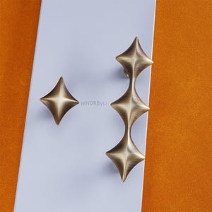 May include: Two brass-toned star-shaped cabinet pulls and a single knob. The pulls have a stacked design, while the knob is a single star. The hardware is set against a white and orange background. The text "WNDRByLi" is visible.