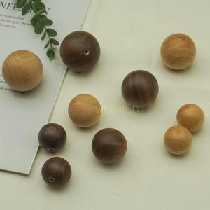 Może przedstawiać: Kolekcja drewnianych kul w różnych odcieniach brązu i beżu. Niektóre kule mają mały otwór. Kule ułożone są na jasnej powierzchni. Obraz zawiera częściowo widoczny tekst.