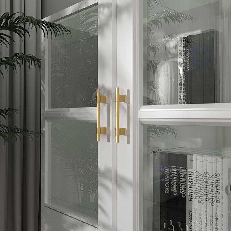 Op de afbeelding: Witte kast met glazen deuren en goudkleurige handgrepen. De kast is gevuld met boeken. De deuren hebben een rasterpatroon. De kast staat voor een raam met een plant.