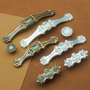 May include: A collection of ornate cabinet hardware in various styles and finishes. The hardware includes handles and knobs with decorative details and inset oval-shaped accents in shades of green and white. The metal finishes include bronze, silver, and gold.