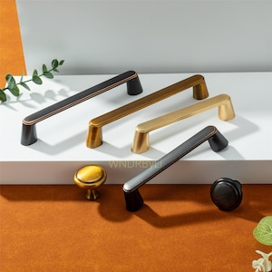 Puede incluir: Una colección de herrajes para armarios en varios acabados metálicos. La imagen muestra varios tiradores de barra en bronce, dorado y negro, junto con dos pomos redondos. Los herrajes se exhiben sobre una superficie blanca con un fondo neutro.