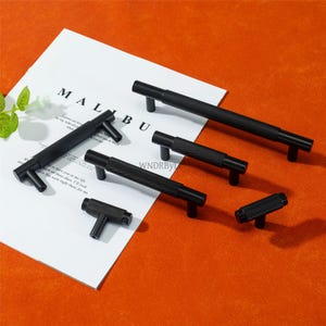 Peut inclure: Plusieurs poignées de meubles noires de différentes tailles, présentées sur du papier blanc avec le mot "MALIBU" imprimé. Les poignées ont une section centrale texturée et des extrémités en forme de T. Le fond est orange uni.