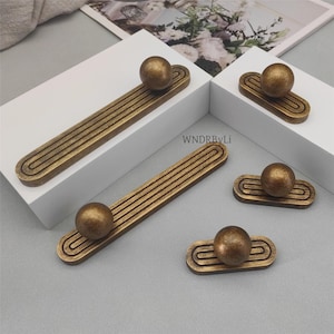 May include: A collection of antique brass cabinet hardware. The hardware includes long, rectangular pulls with a grooved design and a spherical knob at one end, along with smaller, matching knobs. The hardware is displayed on white blocks.