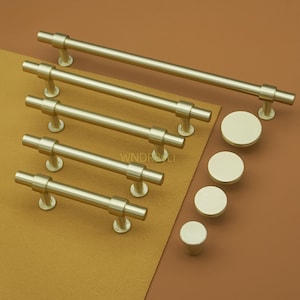 May include: A collection of gold-colored cabinet hardware, including several bar pulls of varying lengths and round knobs. The hardware is arranged on a brown surface, showcasing a modern design aesthetic. The hardware is made of metal.