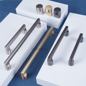 May include: A collection of modern cabinet hardware in various metallic finishes, including silver, gold, and dark gray. The image showcases several handle styles and knob designs, perfect for home decor projects. The text "WNDRByLi" is visible.