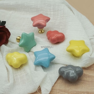 May include: A selection of ceramic cabinet knobs in star and cloud designs. The knobs are in pink, green, yellow, blue, and grey. Each knob has a gold-coloured base. The text "WNDRByLi" is visible.