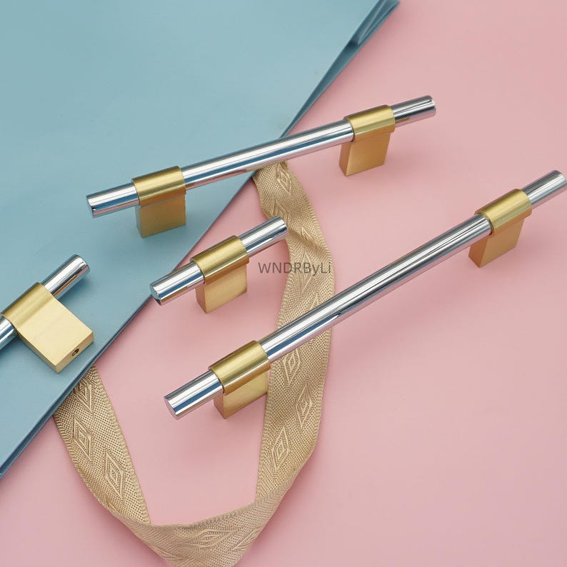 Op de afbeelding: Set van meubelgrepen met een zilver- en goudkleurig kleurenschema. De grepen hebben een zilveren cilindrische staaf met goudkleurige vierkante uiteinden. De afbeelding bevat een decoratief lint en is geplaatst tegen een roze en blauwe achtergrond. De tekst "WNDRBYLi" is zichtbaar.