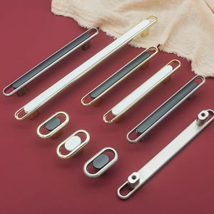 May include: A collection of modern cabinet hardware, including long bar pulls and oval knobs. The pulls feature a mix of black, white, and metallic finishes, with gold and silver accents. The hardware is arranged on a red surface.