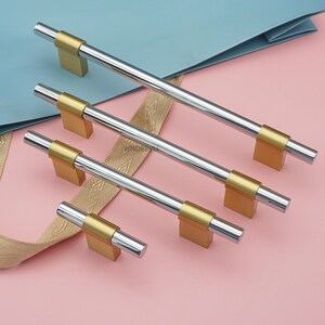 Op de afbeelding: Een set zilver- en goudkleurige kastgrepen in verschillende maten. De grepen hebben een cilindrische zilveren staaf met goudkleurige rechthoekige bases. De achtergrond is een lichtroze oppervlak met een blauw en gouden lint.