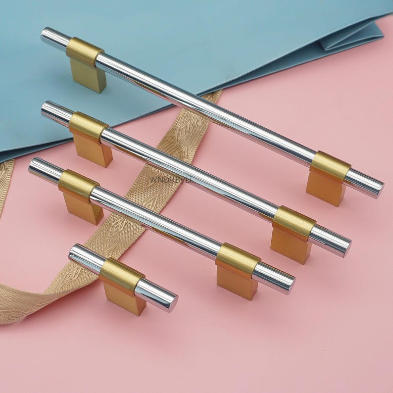 Op de afbeelding: Een set zilver- en goudkleurige kastgrepen in verschillende maten. De grepen hebben een cilindrische zilveren staaf met goudkleurige rechthoekige bases. De achtergrond is een lichtroze oppervlak met een blauw en gouden lint.