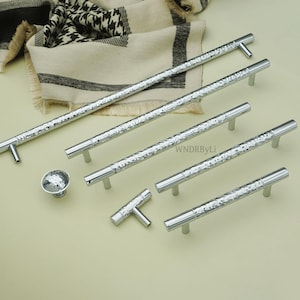 May include: A collection of silver cabinet hardware, including several bar pulls, a knob, and a T-shaped pull. The hardware has a hammered texture and a polished finish. The pulls vary in length.
