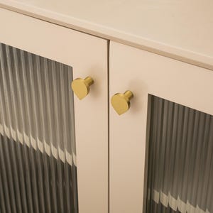 May include: A close-up shot of a cabinet with two doors. The doors have clear, ribbed glass panels and are framed by light beige wood. Each door has a gold-coloured, heart-shaped knob, adding a touch of elegance to the design.