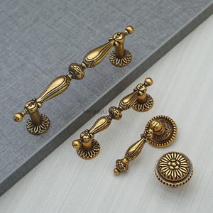 May include: A set of antique-style brass cabinet hardware, including two handles, a smaller handle, and a round knob. The hardware features ornate detailing and a warm, golden finish, suitable for furniture restoration or new builds.