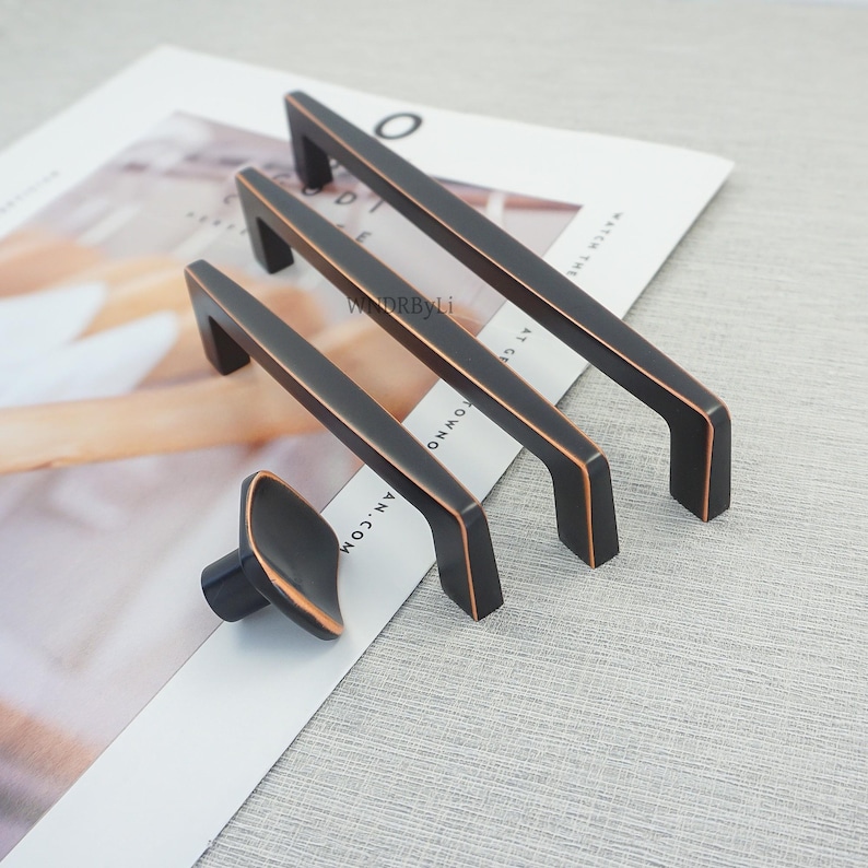 May include: A collection of bronze-toned cabinet handles and a knob. The set includes three rectangular bar pulls and an oval knob. The pulls have a dark finish with copper-coloured edges. The hardware is displayed on a magazine page.