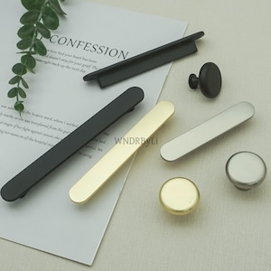 May include: A collection of cabinet hardware in various finishes, including black, gold, and silver. The selection includes bar pulls and round knobs. The hardware is displayed on a white surface with text from a document.