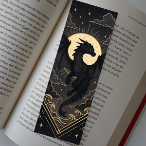 Puede incluir: Marcapáginas de dragón negro con un diseño celestial. El marcapáginas presenta una silueta de dragón sobre un fondo de luna llena, estrellas y nubes. El marcapáginas está sobre un libro abierto.