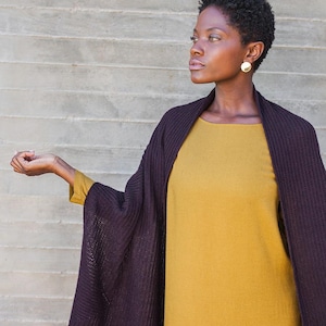 Könnte beinhalten: Eine Frau trägt ein senfgelbes Kleid und einen dunkelvioletten Schal. Der Schal ist über den Schultern und Armen drapiert. Die Frau trägt goldene Ohrringe. Der Hintergrund ist eine graue Betonmauer.