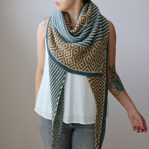 May include: A knitted shawl in shades of brown, teal, and white. The shawl features a geometric diamond pattern and striped sections. The edges are trimmed with a teal border. The shawl is draped over the shoulders.