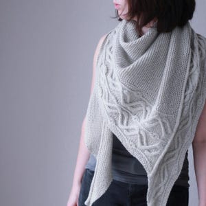May include: A light gray knitted shawl with a cable knit design along the edges. The shawl is draped over the shoulders. The background is a solid light gray.