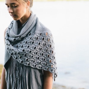 May include: A grey knitted shawl with a distinctive openwork design. The shawl is draped over the shoulders and features a repeating pattern of oval cutouts. The garment is made of a soft, textured yarn, suitable for cooler weather.