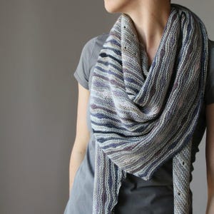 May include: A knitted scarf in shades of grey, purple, and black is draped around a person's neck. The scarf has a striped pattern and a textured appearance. The person is wearing a grey t-shirt. The scarf is a cosy accessory.